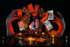 two hands holding playing cards in the water at night with lights on them and candles lit up