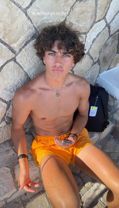 a shirtless young man sitting on the ground next to a stone wall holding his cell phone