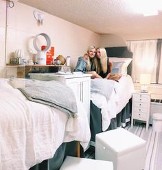 two women sitting on the edge of a bed in a room with white furniture and accessories