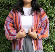 a woman standing in front of a bush wearing a colorful knitted cardigan sweater