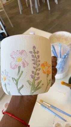 a person holding up a paper cup with flowers painted on it and paintbrushes in the background