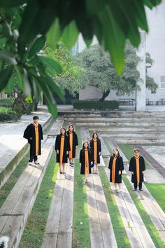 a group of children in graduation gowns walking down a wooden walkway with trees and grass behind them