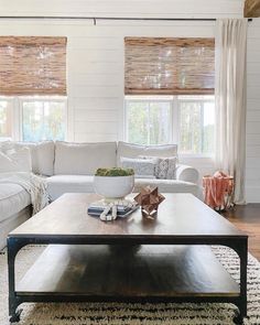 An extra large coffee table takes center stage in this white and wood lounge. A white sofa surrounds the table on stained wooden flooring. A white shiplap accent wall frames white-framed windows with woven roman blinds...   Image: birdsong.design