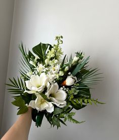 a hand holding a bouquet of white flowers and greenery in front of a wall