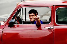 a man in a red car giving the peace sign with his right hand while driving