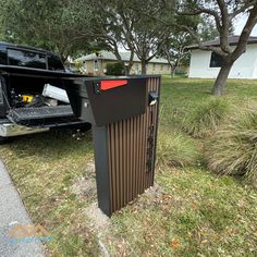 a black trash can sitting in the grass next to a tree and house with it's back door open