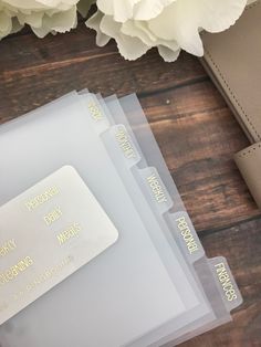 three clear folders with gold lettering on them sitting next to some flowers and a wallet