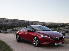 a red car parked on the side of a road near some grass and rocks with mountains in the background