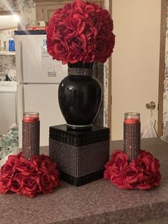 a black vase with red flowers in it on a counter top next to two candles