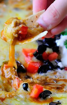 a hand holding a tortilla chip with black olives, tomatoes, peppers and sour cream