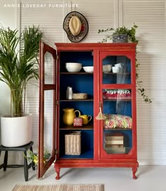 a red china cabinet with blue glass doors and shelves filled with dishes, vases and plants