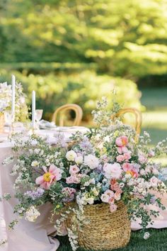 an outdoor table with flowers in a basket and candles on the table, surrounded by greenery