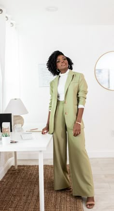 a woman standing in front of a desk with a computer on it and wearing a green suit