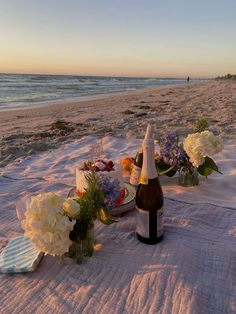 a bottle of wine and some flowers on a blanket at the beach by the ocean