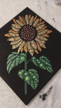 a painting of a sunflower with green leaves on a black square tile that looks like it has been painted