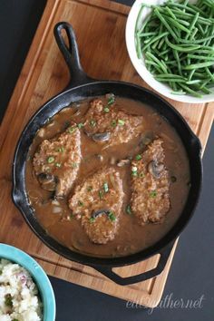 a wooden cutting board topped with green beans and meat covered in gravy next to two bowls of mashed potatoes