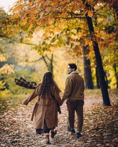 a man and woman holding hands while walking through the woods with leaves on the ground