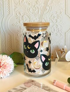 a glass jar with black cats and bats on it sitting next to a pink flower