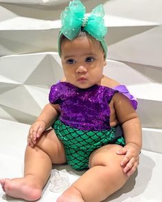 a baby sitting on the ground wearing a purple and green outfit with a bow in it's hair