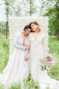 two brides pose for a photo in front of a flower covered backdrop at their wedding