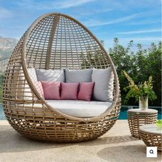 a wicker hanging chair with cushions and pillows on the ground next to a pool