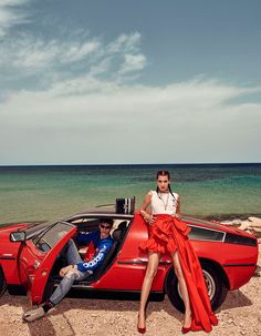 two people sitting on the hood of a red sports car near the ocean with their feet in the open door