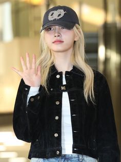 a woman with long blonde hair wearing a black jacket and hat holding her hand up in the air