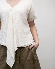 a woman wearing a white top with a tie around her neck and short sleeves, standing in front of a wall