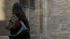 a woman carrying a black purse walking down the street in front of a stone building