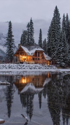 a log cabin is lit up at night by the water's edge with snow on the ground