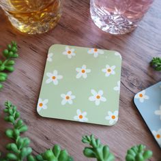 a coaster with daisies on it next to two glasses and green plant stems in the foreground