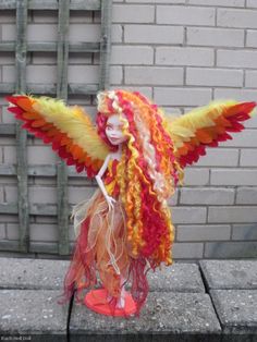 a doll is dressed up like a fairy with orange, yellow and red feathers on her head