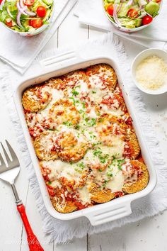 a casserole dish with meat, cheese and vegetables next to salad on a white table