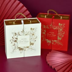 two red and gold gift bags next to each other on a purple surface with fan