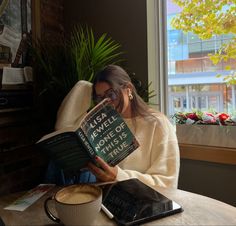 a woman sitting in a chair reading a book next to a cup of cappuccino