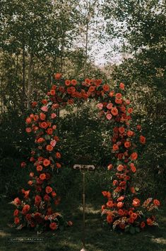 an arch made out of red flowers in the middle of a field with trees behind it