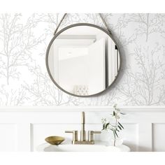 a bathroom sink under a round mirror next to a white wall with trees on it