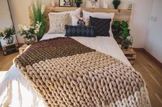 a bed covered in lots of pillows on top of a wooden floor next to potted plants