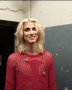 a woman with blonde hair standing in front of a gray wall wearing a red sweater