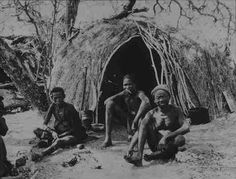 an old black and white photo of people sitting in front of a hut with grass roof