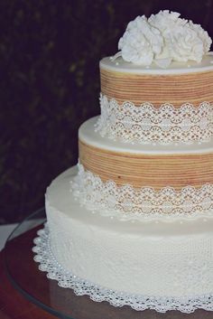 a three tiered wedding cake with lace and flowers on the top is sitting on a table