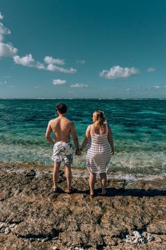 a man and woman holding hands while walking on the rocks near the water's edge