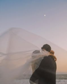 a man and woman standing on top of a beach next to the ocean under a veil