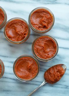 six jars filled with red sauce sitting on top of a marble counter next to spoons