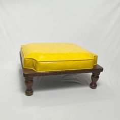 a yellow ottoman with wooden legs on a white background in front of a plain backdrop