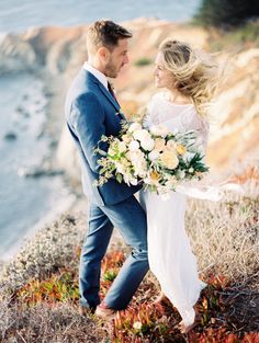 a bride and groom are walking on the cliff by the ocean with flowers in their hands