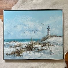 a painting of a lighthouse on the beach with seagulls flying over it and sand dunes in the foreground