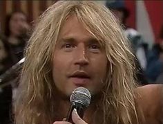 a man with long blonde hair holding a microphone in front of his face and looking at the camera