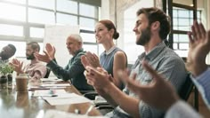 a group of people sitting around a conference table clapping hands in front of the camera