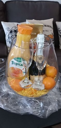 two wine glasses and bottles in a glass bowl with oranges on the table next to it